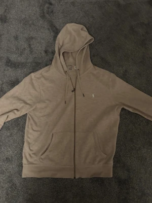 Beige hoodie från Polo Ralph Lauren - Snygg beige hoodie från Polo Ralph Lauren med dragkedja och huva med snörning. Tröjan har en klassisk passform, långa ärmar och en broderad logga på bröstet. Tillverkad i mjuk bomull, perfekt för chill dagar. Fickor framtill för extra casual vibe.