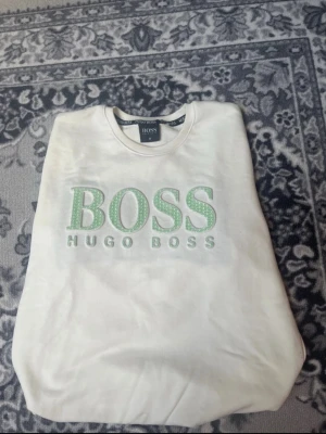 Vit sweatshirt från Hugo Boss M - Vit sweatshirt från Hugo Boss i storlek M med stort grönt BOSS-tryck och broderad logga på bröstet. Tröjan har rund hals och långa ärmar, perfekt för dig som gillar stilrena och klassiska märkesplagg. Mjuk bomullskänsla och relaxed fit.