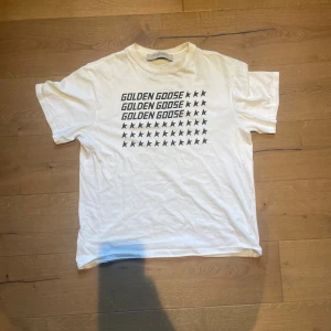 Vit t-shirt från Golden Goose - Vit t-shirt från Golden Goose med svart tryck framtill där det står 'GOLDEN GOOSE' och flera svarta stjärnor. Klassisk passform och rund halsringning. Tillverkad i mjuk bomull, perfekt för en clean och stilren look.