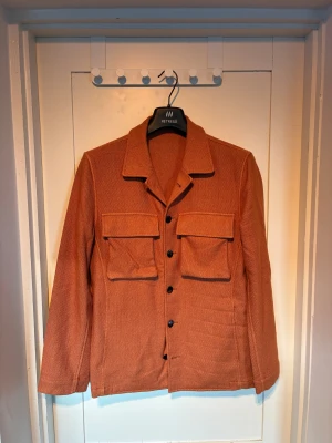 Rostfärgad overshirt från Lardini - Overshirt från Lardini i en snygg rostbrun ton, tillverkad i Italien. Jackan har två stora bröstfickor med lock, svarta knappar framtill och klassisk krage. Materialet är grovt vävt och ger en exklusiv känsla. Perfekt för lager-på-lager-stil. Nypris 6.000, mitt pris 1750