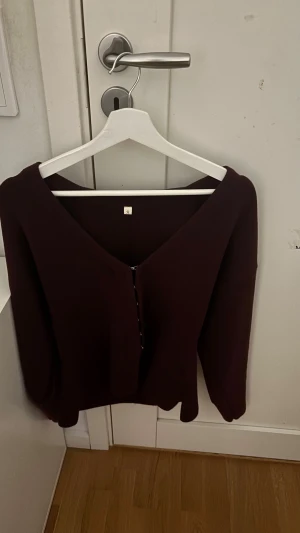 Vinröd kofta med v-ringning, stl S - Vinröd kofta med djup v-ringning och dekorativa knappar framtill. Modellen har långärmade ärmar och en loose passform som ger en chill vibe. Perfekt för lager-på-lager och att slänga över en t-shirt. Materialet känns mjukt och är troligtvis stickat.