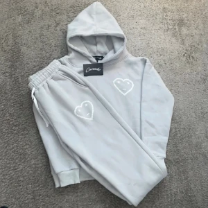 Ljusgrå hoodie med hjärtmotiv från Carsiko - Säljer en ljusgrå hoodie från Carsiko med huva och ribbade muddar. På bröstet finns ett broderat vitt hjärta med ett ansikte och små stjärnor. Hoodien är i mjuk bomull och har en relaxed passform, perfekt för chill dagar.