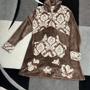 Brun mönstrad velourklänning - Unik brun velourklänning med lång ärm och hög krage. Klänningen har ett dekorativt vitt mönster på bröstet, fickan, ärmsluten och nedre delen. Mjuk och skön känsla, perfekt för dig som gillar retrovibbar och statement-plagg.