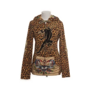 Leopard hoodie från Christian Audigier - Unik hoodie med leopardmönster från Christian Audigier. Tröjan har huva med snörning, coolt svart leopard framtill och detaljerad broderad ficka med cheetah motiv. Baksidan har en stor silverkrona. Materialet är bomull och polyester, med långärmad passform.