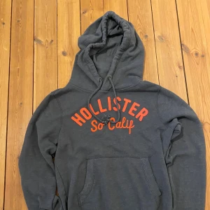 Holister Hoodie - En hoodie med perfekt passform och premiumkänsla. Inspirerad av klassisk Hollister-stil – avslappnad men stilren. Tillverkad i mjuk bomullsblandning med borstad insida för extra komfort. Ribbstickade muddar och justerbar huva med snörning ger en lyxig street-look. Passform: Regular-fit (sitter snyggt men inte för tight). Storlek: S Design: Enkel, ren logga på bröstet eller ton-i-ton broderi.