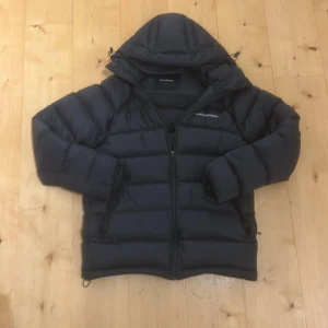 Svart Peak Performance pufferjacka M - Säljer en svart pufferjacka från Peak Performance i storlek M. Jackan har huva, dragkedja framtill och broderad logga på bröstet och armen. Den är vadderad och har en clean look. Perfekt för kalla dagar och dåligt väder.