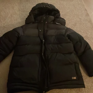 Svart pufferjacka från Lexington XS - Säljer en svart pufferjacka från Lexington i storlek XS. Jackan har huva, grov dragkedja med Lexington-logga och quiltad design. Perfekt för kalla dagar och har en clean look som funkar till allt. Fickor på sidorna och snygg Lexington-märke nedtill. Kostar 5500kr ny.