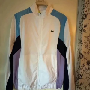 Lacoste  vindjacka - En riktigt snygg vindjacka från Lacoste köpt på nätet för cirka 1 år sen. Jag använder inte den då jag har bytt stil. Den är använd cirka 6-7 gånger. Den är helt ny och ligger bara nu i garderoben. 