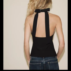 Svart halterneck topp med öppen rygg - Säljer en svart halterneck topp med breda axelband och snygg v-ringning framtill. Toppen har öppen rygg och en figurnära passform som ger en clean och modern look. Perfekt att styla med jeans eller kjol för en trendig outfit.