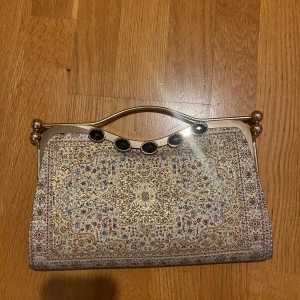 Glittrig kuvertväska med orientaliskt mönster - Säljer en unik kuvertväska med orientaliskt, glittrigt mönster i guld, beige och inslag av rött och blått. Väskan har ett metallhandtag med svarta dekorstenar och en lyxig känsla. Perfekt för dig som vill sticka ut med en elegant accessoar.