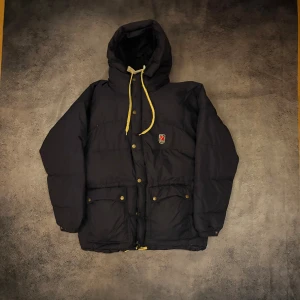 Mörkblå dunjacka från Fjällräven M - Säljer en mörkblå dunjacka från Fjällräven i storlek M. Jackan har huva med gula snören, tryckknappar och dragkedja framtill samt två stora fickor med knappstängning. Klassisk Fjällräven-logga på bröstet. Perfekt för kalla dagar.