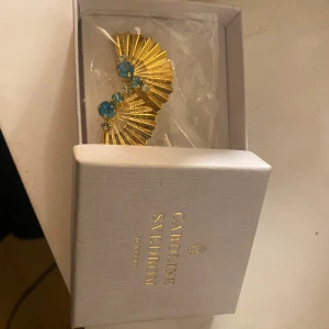 Guldörhängen med blå stenar Caroline Svedbom - Unika örhängen från Caroline Svedbom i form av solfjädrar i guldton med blå gnistrande stenar. De har en cool, veckad design och detaljerade mönster på metallen. Perfekta för dig som vill sticka ut med ett statement-smycke.