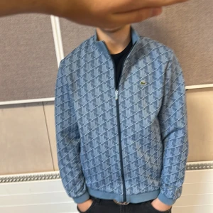 Blå mönstrad bomberjacka från Lacoste - Säljer en blå bomberjacka från Lacoste med vitt geometriskt mönster och klassisk krokodillogga på bröstet. Jackan har ribbade muddar, dragkedja framtill och står ut med sin coola design. Perfekt för dig som vill sticka ut med stil.