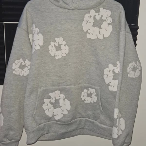 Denim Tears Hoodie - Säljer en grå hoodie från Denim Tears i storlek S med vita blommor tryckta över hela tröjan. Hoodien har huva, magficka och en relaxed passform. Perfekt för dig som vill ha en lite relaxed stil.