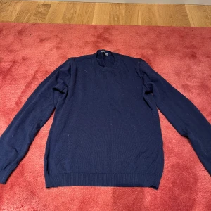 Mörkblå stickad tröja med mockadetaljer från Nikolaj - Snygg mörkblå stickad tröja med rund hals och långa ärmar. På baksidan finns bruna mockalappar på armbågarna med broderad detalj, vilket ger en cool och klassisk touch. Perfekt för dig som gillar stilrena och tidlösa plagg.