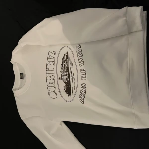 Corteiz sweatshirt - Vit sweatshirt från Corteiz med tryck 'RULZ THE WORLD' och grafiskt motiv på bröstet. Tröjan har rund hals, ribbade muddar och lång ärm. Perfekt för dig som gillar streetwear och vill sticka ut med ett snyggt statement. Storlek M men skulle säga mer som S. 