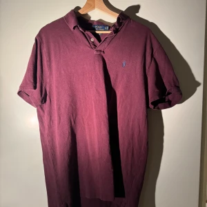 Vinröd pikétröja Polo Ralph Lauren XL - Snygg vinröd pikétröja från Polo Ralph Lauren i storlek XL. Tröjan har klassisk krage, korta ärmar och den ikoniska broderade loggan på bröstet. Modellen är custom slim fit och är gjord i mjuk bomull som känns skön mot huden.