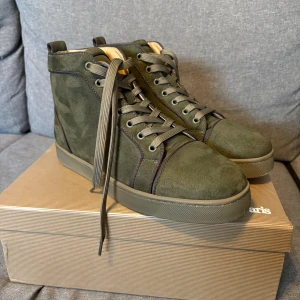 Olivgröna sneakers från Christian Louboutin - Snygga olivgröna high-top sneakers från Christian Louboutin i mjuk mocka med signaturröd sula. Skorna har snörning, rund tå och diskret broderad logga på sidan. Perfekt för dig som vill sticka ut med exklusiva detaljer och streetstyle-vibe.