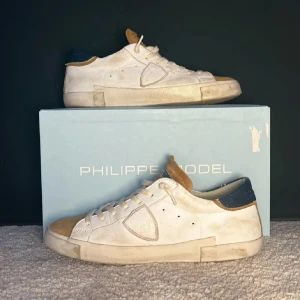 Philippe Model vita sneakers med mockadetaljer - Snygga vita sneakers från Philippe Model med beige mockadetaljer på tå och plös samt mörkblå häl. Klassisk låg modell med rund tå, snörning och platt sula. Perfekta för dig som gillar stilrena och exklusiva sneakers med coola kontraster.