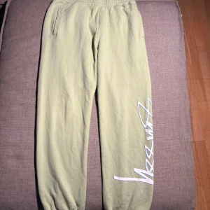 Stüssy ljusgröna mjukisbyxor - Ljusgröna mjukisbyxor från Stüssy med elastisk midja och muddar vid benslut. Byxorna har fickor på sidorna och en bakficka, samt Stüssy-logga i vitt med blå kontur längs ena benet. Perfekta för chill eller streetwear.