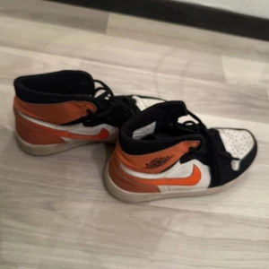 Nike Air Jordan 1 orange och svart - Säljer ett par Nike Air Jordan 1 med högt skaft i en riktigt snygg färgkombo av orange, svart och vitt. Skorna har klassisk snörning, perforerad tåbox och ikoniska swoosh-loggan på sidan. Perfekta för dig som vill sticka ut med dina sneakers.