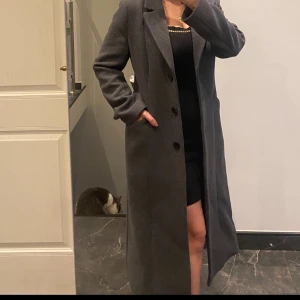 Mörkgrå lång kappa med knappar - Snygg mörkgrå kappa med klassisk krage och tre svarta knappar framtill. Modellen är lång och har en rak passform med diskreta fickor på sidorna. Perfekt för dig som gillar stilrena och tidlösa plagg. Materialet känns som ull och kappan har långa ärmar.