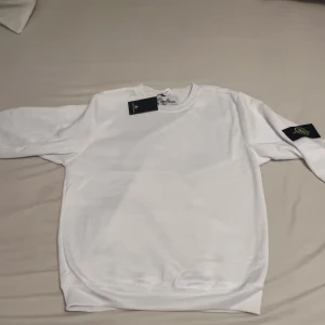 Vit sweatshirt från Stone Island XL - Vit sweatshirt från Stone Island i storlek XL. Klassisk rund hals och långärmad modell med den ikoniska Stone Island-patchen på vänster ärm. Tröjan är gjord i mjuk bomull och har en clean look som passar perfekt till jeans eller cargo.
