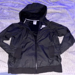 Svart Nike vindjacka med huva - Svart vindjacka från Nike med vit logga på bröstet. Jackan har huva, hel dragkedja framtill och ribbade muddar vid ärmslut och nederkant. Tillverkad i lätt polyester med meshfoder för extra komfort. Perfekt för dig som gillar sportig stil.