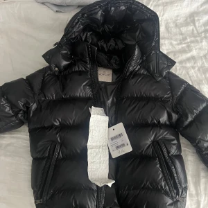 Svart Moncler pufferjacka med huva (Kvitto finns) - Säljer en svart pufferjacka från Moncler med glansig finish och huva. Jackan har två sidofickor med dragkedja, klassisk Moncler-logga på ärmen och är fylld med vitt gåsdun. Perfekt för kalla dagar och har en snygg, modern look.