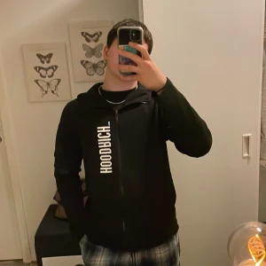 Svart hoodie från Hoodrich med tryck - Svart hoodie från Hoodrich med vit text på bröstet, ryggen och huvan. Snygg streetwear-stil med dragkedja framtill och broderad FNTS™ på ärmen. Klassisk passform och huva. Perfekt för dig som gillar hoodrich 