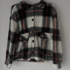 Rutig teddyjacka med knappar - Mysig teddyjacka i rutig design med svart, vit och rosa färg. Jackan har krage, långa ärmar och stora knappar framtill. Två fickor med lock och knappdetalj på framsidan. Perfekt för dig som gillar street och chill stil.