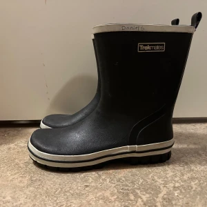 Svarta stövlar från Trekmates strl: 37 - Svarta stövlar från Trekmates med vit kant upptill och längs sulan. Grov sula för extra grepp och klassisk rund tå. Tillverkade i syntetmaterial som tål väta, perfekta för regniga dagar. Enkla att dra på med två flikar upptill. Står namn på det vita.