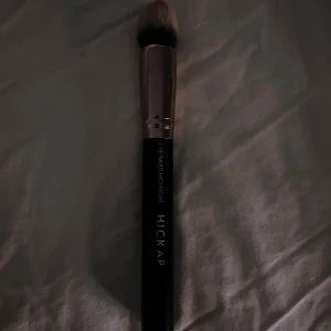 Hickap 119 Multi Highlight borste - Snygg sminkborste från Hickap, modell 119 Multi Highlight. Borsten har svart skaft med silverfärgad metall och täta, mjuka borststrån i beige och brunt. Perfekt för att applicera highlighter eller puder med precision.