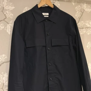 Svart overshirt från Massimo Dutti - Stilren svart overshirt från Massimo Dutti Studio med klassisk krage. Skjortan har knäppning framtill och är långärmad. Materialet är slätt och skönt, perfekt för dig som gillar minimalistisk design. Perfekt nu till hösten och kallar väder, storlek s men skulle säga att den passar M! 🍂🍁😊