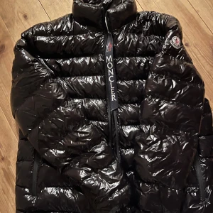 Svart pufferjacka från Moncler - Säljer en svart pufferjacka från Moncler med glansig finish och hög krage. Jackan har tydliga Moncler-loggor, dragkedja framtill och två sidofickor med dragkedja. Insidan har en cool serietidningsetikett och jackan är perfekt för kalla dagar.