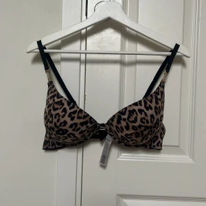 Leopardmönstrat Bh - Snygg Bh med leopardmönster i beige och svart, svarta axelband och glittrande strassdetaljer i guld vid banden och mellan kuporna. Materialet är mjukt och glansigt, Perfekt för dig som vill sticka ut med djurprint och bling. Med push up.