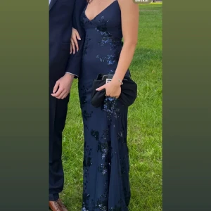 Marinblå balklänning med snörad rygg - Säljer min marinblå balklänning med glittriga detaljer och blommigt mönster. Klänningen har smala axelband, v- rigning och öppen rygg med  snörning. 