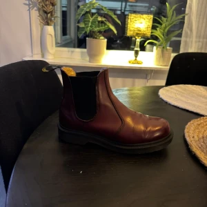Vinröda Chelsea boots i skinn - Säljer ett par stilrena Chelsea boots i vinrött skinn med elastiska svarta paneler på sidorna och en robust svart platt sula. Skorna har rund tå och klassisk dragflik bak för enkel påtagning. Perfekt för dig som gillar tidlös och edgy stil.