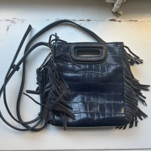 Blå liten axelväska med fransar från Maje - Liten blå axelväska från Maje i skinn med fransar längs sidorna. Väskan har både handtag och axelrem, samt snygga guldfärgade detaljer. Perfekt storlek för det viktigaste och levereras med dustbag.