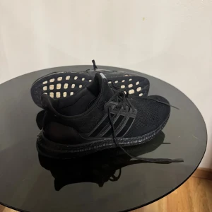 Svarta Adidas Ultraboost sneakers - Säljer ett par svarta Adidas Ultraboost sneakers med stickad ovandel och platt sula. Skorna har snörning och en diskret Adidas-logga på plösen. Boost-sulan ger grym komfort och respons, perfekt för dig som gillar att röra på dig. Snygg, helsvart look med mesh och syntetdetaljer.