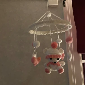 Virkad baby mobil med björn och hjärtan - Charmig handvirkad baby mobil i vitt och rosa med söt björn, hjärtan och bollar. Perfekt för barnrummet eller som present. Enkel att hänga upp med medföljande ring. Skapar en mysig och lekfull atmosfär.