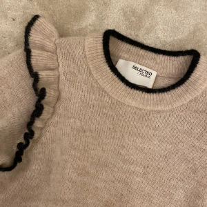 Beige stickad tröja med volang Selected Femme - Superfin beige stickad tröja från Selected Femme med svarta kontrastkanter och volangdetaljer på axlarna. Köpt för 1000kr och nästan helt oanvänd. Tröjan har rund halsringning, långa ärmar och ribbade muddar. Perfekt för dig som gillar en söt och trendig look.