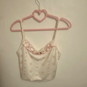 Söt blommig linnetopp med volang - Supersöt linnetopp i ljus cremefärg med små rosa blommor och rosetter. Toppen har tunna axelband, volangkant med spets och en liten tygros framtill. Perfekt för dig som gillar romantisk och girly stil. Materialet känns mjukt och lätt.