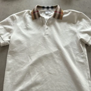 Vit Burberry pikétröja med rutig krage - Vit pikétröja från Burberry med klassisk rutig krage i beige, svart och rött. Tröjan har korta ärmar, knappar vid halsen och är tillverkad i mjuk bomull. Perfekt för dig som gillar stilrena och ikoniska detaljer.