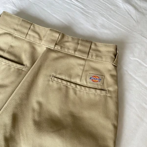 Beige Dickies 874 byxor W29/L30 - Klassiska beige Dickies 874 Original Fit byxor med raka ben och normal passform. Byxorna har hällor för bälte, två bakfickor och Dickies-logga ovanför ena fickan. Tillverkade i slitstarkt material som passar perfekt till en streetwear-look. Finns ett litet streck på insidan låret, skriv privat för bild.
