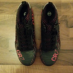 Svarta CR7 fotbollsskor med ljus röda detaljer - Säljer ett par svarta CR7 fotbollsskor med ljus röda och vita detaljer. Perfekt skick utan skador eller liknande och funkar jättebra med grepp och snabbhet. Fråga om ni undrar något eller om ni är intresserade😁!