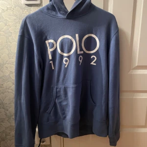 Blå hoodie Polo Ralph Lauren M - Snygg blå hoodie från Polo Ralph Lauren med stor vit print 'POLO 1992' på bröstet. Tröjan har huva, känguruficka och långa ärmar. Perfekt för dig som gillar klassisk streetstyle och vill ha en chill look.