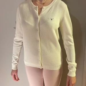 Vit kofta från Gant i bomull - Klassisk vit kofta från Gant med rund halsringning och diskret broderad logga på bröstet. Koftan har knäppning framtill, ribbade muddar och är tillverkad i mjuk bomull. Perfekt för lager-på-lager och enkel att matcha med allt.