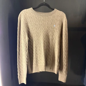 Beige kabelstickad tröja från Morris - Kabelstickad beige tröja från Morris med rund halsringning och diskret broderad logga på bröstet. Tröjan har lång ärm och klassisk passform, perfekt för lager-på-lager-stil under höst och vinter.