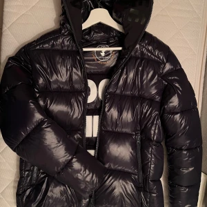 Mörkblå pufferjacka från Save The Duck - Säljer en mörkblå pufferjacka från Save The Duck med glansig finish och stor vit text '100% Animal Free' på insidan. Jackan har huva, två sidofickor med dragkedja och är tillverkad i syntetmaterial. Perfekt för kalla dagar nu till vintern ❄️ 